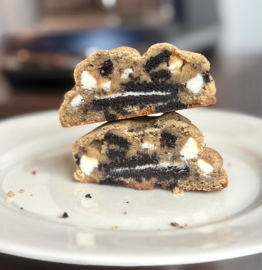 Two halves of a cookies-and-cream cookie stacked on a white plate, packed with white chocolate chunks and a whole sandwich cookie.